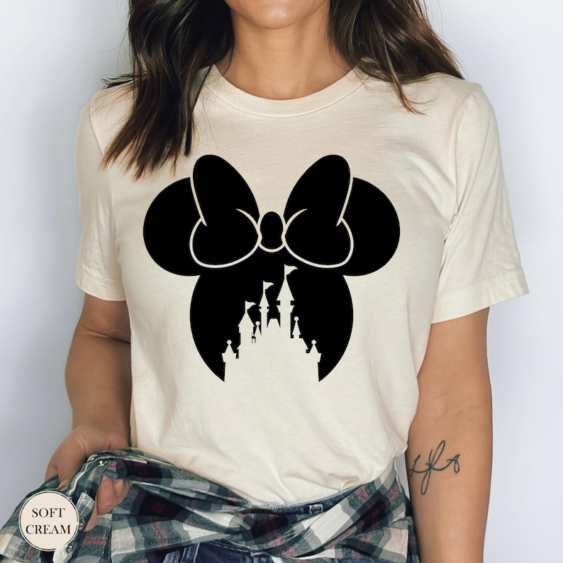 May include: Cream-colored t-shirt featuring a black silhouette of a bow with a castle design inside. The text "SOFT CREAM" is printed on the bottom left of the shirt. The shirt is made of a soft material.