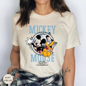 May include: Soft cream-colored t-shirt featuring a graphic of Mickey Mouse and Pluto. The design includes the words "Mickey Mouse" in light blue, with a circular image of the characters. The shirt has short sleeves.