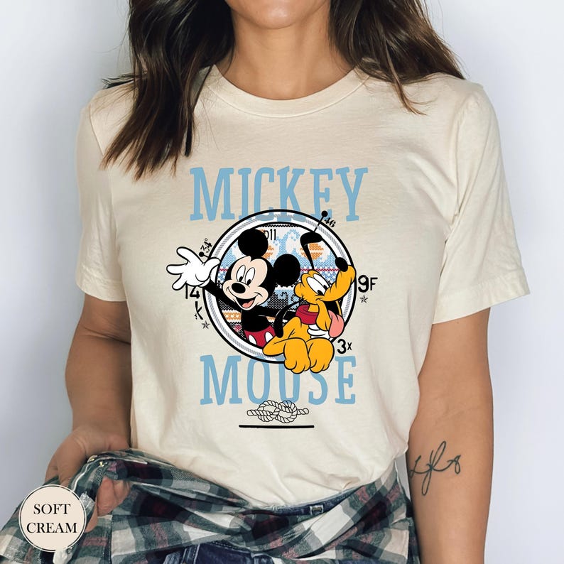 May include: Soft cream-colored t-shirt featuring a graphic of Mickey Mouse and Pluto. The design includes the words "Mickey Mouse" in light blue, with a circular image of the characters. The shirt has short sleeves.