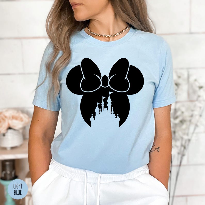 May include: Light blue t-shirt featuring a black silhouette of a bow with a castle design. The shirt has short sleeves and a crew neck. The words "LIGHT BLUE" are printed on a small label in the bottom left corner.