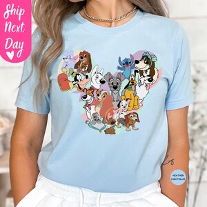 May include: Light blue t-shirt featuring a heart-shaped design composed of various cartoon dog characters. The shirt has short sleeves and a crew neck. The text "Ship Next Day" is on the left side of the shirt.