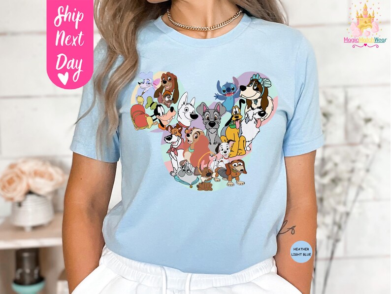 May include: Light blue t-shirt featuring a heart-shaped design composed of various cartoon dog characters. The shirt has short sleeves and a crew neck. The text "Ship Next Day" is on the left side of the shirt.