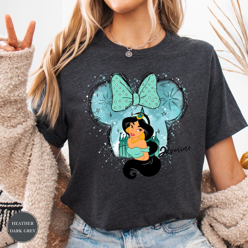 Puede incluir: Camiseta gris oscuro con una ilustraci&oacute;n de dibujos animados de la Princesa Jasmine dentro de la forma de una cabeza de Mickey Mouse. El dise&ntilde;o incluye un lazo turquesa, un castillo y el nombre "Jasmine". La camiseta tiene el texto "Heather Dark Grey".