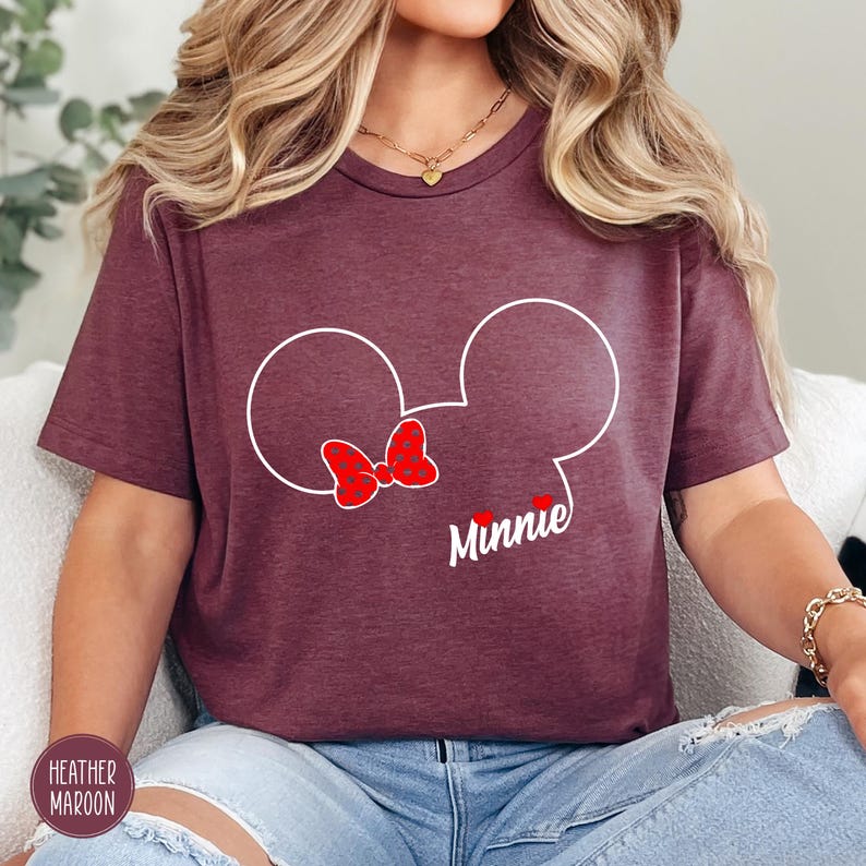 May include: Heather maroon t-shirt featuring a white outline of Minnie Mouse's head with a red bow and the name "Minnie" in script. The shirt is made of a soft material and is suitable for casual wear.