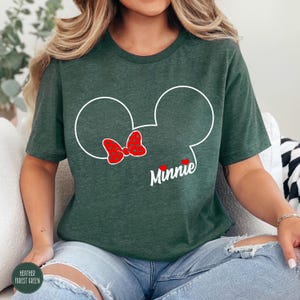 May include: A dark green t-shirt featuring a white outline of Minnie Mouse's head with a red polka-dot bow. The name "Minnie" is written in white script below the bow. The shirt is made of a soft material.