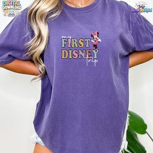 May include: A purple short-sleeve t-shirt featuring a graphic print with the text "my FIRST DISNEY trip" in multi-coloured block letters and a small illustration of Minnie Mouse wearing a pink dress and bow positioned above the word trip.