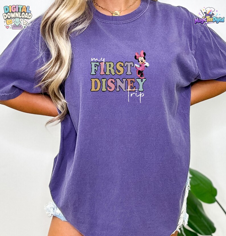 May include: A purple short-sleeve t-shirt featuring a graphic print with the text "my FIRST DISNEY trip" in multi-coloured block letters and a small illustration of Minnie Mouse wearing a pink dress and bow positioned above the word trip.