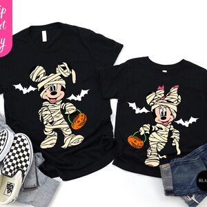 May include: Black t-shirts featuring Mickey and Minnie Mouse dressed as mummies for Halloween. Each character holds a pumpkin-shaped candy bucket. White bats are printed above the characters. The text "Ship Next Day" is on the left.