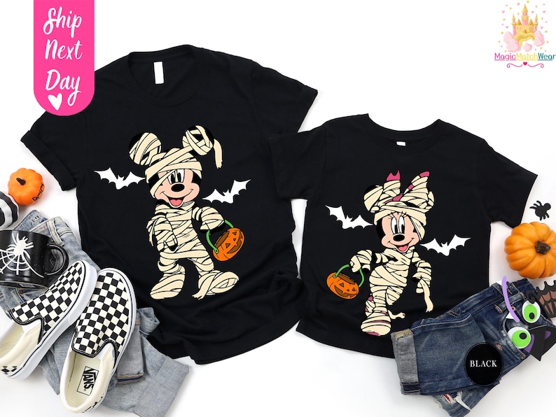 May include: Black t-shirts featuring Mickey and Minnie Mouse dressed as mummies for Halloween. Each character holds a pumpkin-shaped candy bucket. White bats are printed above the characters. The text "Ship Next Day" is on the left.