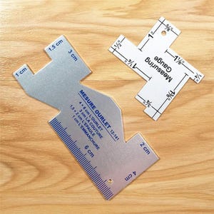 May include: A silver-colored metal sewing gauge with blue markings for 1 cm, 1.5 cm, 2 cm, 3 cm, 4 cm, and 6 cm measurements, alongside a white plastic measuring gauge with black markings for 1/4 inch, 1/2 inch, 5/8 inch, 1 inch, 1 1/8 inch, and 1 3/8 inch.