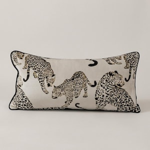 Puede incluir: Cojín rectangular con fondo beige y un estampado repetido de leopardos negros y marrones. El cojín tiene un borde negro.