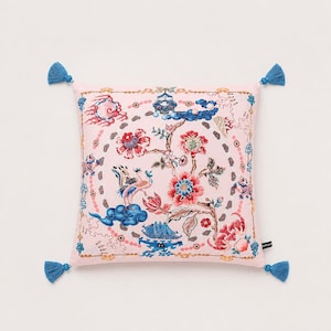 May include: A square decorative pillow with a pink background and a floral and bird pattern in red, blue, and gold. The pillow has four blue tassels, one on each corner. The design is reminiscent of traditional Asian art.
