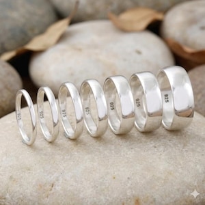 May include: Six silver rings of different widths are displayed on a light-coloured stone. Each ring is stamped with "925". The rings have a polished, reflective finish and are arranged in a row, showing their size variations. The rings are made of silver.