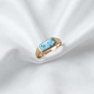 May include: A gold-toned signet ring featuring a rectangular, light blue stone with white marbled patterns. The ring has a smooth, polished metal band and a flat, bezel-set stone inlay that sits flush within the top surface of the ring setting.