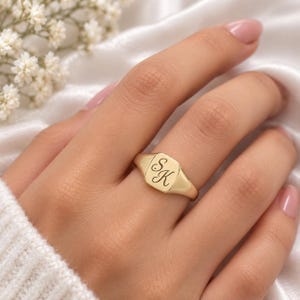 May include: A gold signet ring featuring a smooth, polished surface with the engraved letters SK in a cursive script font. The ring has a flat, rectangular face with rounded corners and a tapered band, showcasing a classic, metallic finish.