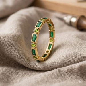 Könnte beinhalten: Ein goldener Ring mit rechteckigen und runden grünen Edelsteinen. Der Ring ist in einem goldenen Band gefasst und liegt auf einem neutralfarbenen Stoff. Die Edelsteine haben eine leuchtend grüne Farbe.
