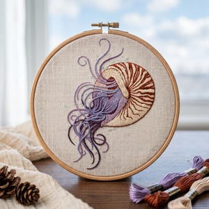 May include: A circular wooden embroidery hoop containing a fabric piece with a hand-stitched nautilus shell and flowing tentacles. The shell features brown and cream stripes, while the tentacles are rendered in shades of purple and lavender thread.
