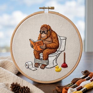 May include: An embroidery hoop containing a stitched image of a brown orangutan wearing black glasses, sitting on a white toilet while reading an orange book titled TOILET JOKES. A roll of white toilet paper, a red plunger, and a yellow rubber duck are included.