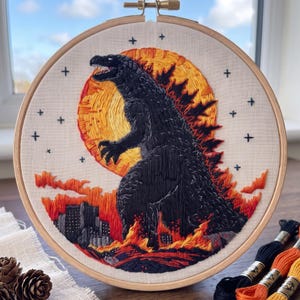 May include: A circular embroidery hoop containing a hand-stitched design of a large black monster standing amidst orange and red flames. A bright yellow and orange sun sits behind the creature, with small black cross-shaped stars in the sky.
