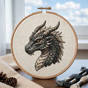Könnte beinhalten: Ein runder Stickrahmen aus Holz mit einem Stoffeinsatz, der einen detaillierten, mit Faden gestickten Drachenkopf zeigt. Der Drache hat dunkelgraue und schwarze Schuppen, eine strukturierte Mähne und ein sichtbares orangefarbenes Auge.