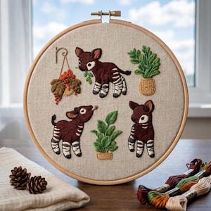 May include: A circular wooden embroidery hoop containing a beige fabric panel with hand-stitched designs. The embroidery features three okapis with dark brown bodies and white striped legs, alongside two potted green plants and a hanging basket.