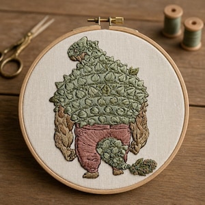 Puede incluir: Arte de aro bordado que presenta una criatura verde y marrón con forma de dinosaurio con una espalda puntiaguda, que lleva pantalones rosas. La obra de arte está sobre una tela color crema, enmarcada por un aro de madera. Tijeras y carretes de hilo en el fondo.