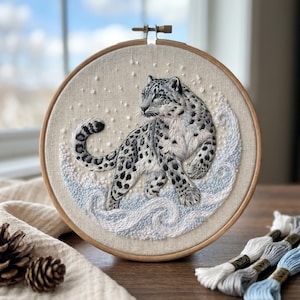 May include: A circular wooden embroidery hoop containing a detailed needlework piece of a snow leopard. The leopard has white and grey fur with black spots, positioned amidst swirling light blue and white embroidered waves and falling snow.