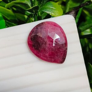Puede incluir: Una piedra preciosa en forma de pera con una superficie facetada, que muestra un degradado de color magenta intenso y rojo oscuro. La piedra presenta un acabado pulido con inclusiones internas visibles y una sección superior reflectante.