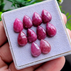 Puede incluir: Un conjunto de nueve piedras preciosas de rubí facetadas en forma de pera con un tono rojo rosado intenso. Las piedras están dispuestas en un grupo sobre un inserto de espuma gris claro texturizado dentro de una caja de presentación cuadrada blanca.