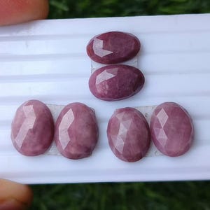 May include: Six oval-shaped, faceted gemstones with a deep mauve and dusty rose colour palette. The stones are arranged on a white, ridged display card, showcasing a polished surface with geometric facets that reflect light across their smooth tops.