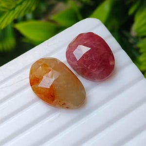Puede incluir: Dos piedras preciosas facetadas de forma ovalada. Una piedra es de color ámbar anaranjado translúcido con inclusiones internas visibles. La segunda piedra es de un color rojo frambuesa profundo y opaco con una superficie pulida y facetas geométricas.