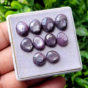 Peut inclure: Une collection de neuf pierres précieuses violettes à facettes de forme ovale, disposées dans une boîte de présentation blanche. Chaque pierre présente une surface polie avec des inclusions internes visibles et une teinte violette profonde.