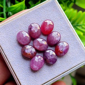 May include: A collection of eight oval-shaped, faceted rubies arranged in a cluster on a light grey, textured display pad. Each gemstone exhibits a deep pinkish-red hue with visible internal inclusions and a polished, multi-faceted surface.