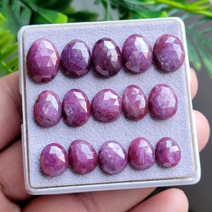 May include: A collection of fifteen oval-shaped, faceted ruby gemstones arranged in three rows of five within a square white display box. Each gemstone features a deep magenta-red hue with a polished, multi-faceted surface that reflects light.