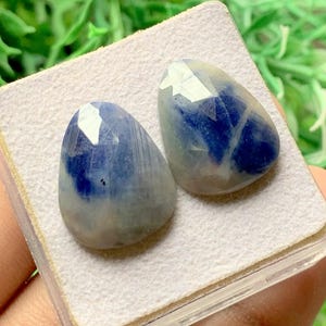 Peut inclure: Deux pierres précieuses facettées en forme de poire avec une surface lisse et polie. Les pierres présentent un dégradé de couleurs distinct, passant d'un bleu profond et saturé au sommet à une base blanc laiteux translucide avec des inclusions.