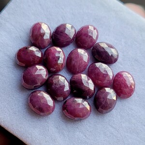 Puede incluir: Una colección de piedras preciosas de rubí ovaladas y facetadas. Los rubíes son de un color rojo intenso, con un ligero matiz púrpura. Las facetas capturan la luz, creando un efecto brillante. Las piedras están dispuestas sobre una superficie gris claro.