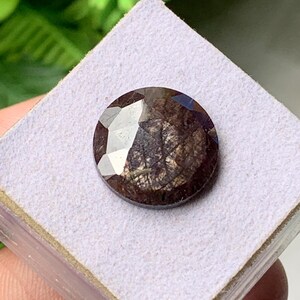 May include: A round, faceted gemstone with a deep, dark brown and black colour palette. The stone features a complex, textured surface with visible internal inclusions and a polished, reflective finish, showcasing a mix of light and dark tones.