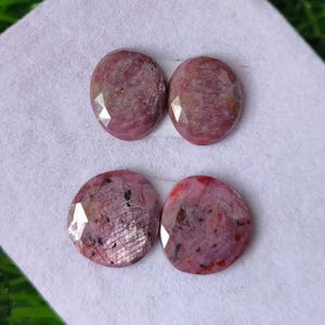 May include: Four oval-shaped, faceted rubies with a deep pinkish-red hue. Each gemstone displays a polished surface with visible internal inclusions and natural patterns, arranged in two pairs on a light-coloured, textured fabric surface.