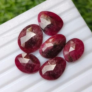 Peut inclure: Six rubis facettés de forme ovale aux teintes rouge profond avec des inclusions internes visibles. Chaque pierre précieuse présente une surface polie avec des coupes géométriques qui reflètent la lumière, disposées sur une surface blanche striée.