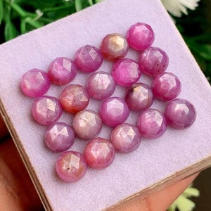 Può includere: Una collezione di diciassette rubini rosa rotondi e sfaccettati disposti a griglia. Ogni gemma presenta un taglio brillante con molteplici sfaccettature triangolari che riflettono la luce, mostrando varie tonalità di magenta e rosa vibrante.