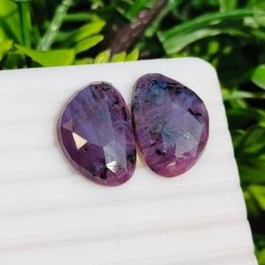 May include: Two faceted, semi-oval gemstones with a deep purple and magenta hue. The stones feature irregular, dark inclusions throughout their surfaces, creating a natural, mottled appearance with reflective, geometric cut facets across the top.
