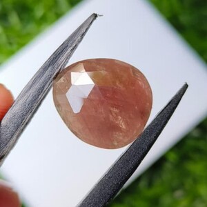 Puede incluir: Una piedra preciosa facetada de forma ovalada con un tono rosa melocotón cálido. La piedra presenta una superficie pulida con múltiples facetas geométricas que reflejan la luz, sostenida entre las dos puntas de unas pinzas de color gris oscuro.