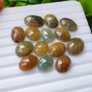 May include: A collection of thirteen oval-shaped, faceted gemstones in various shades of brown, amber, and muted green. Each stone features a polished, multi-faceted surface that reflects light, arranged in a cluster on a white ridged surface.