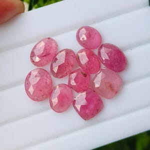 May include: A collection of nine faceted pink gemstones with oval, round, and cushion shapes. Each stone displays a translucent quality with internal inclusions and a polished surface, reflecting light across their geometric cut facets.