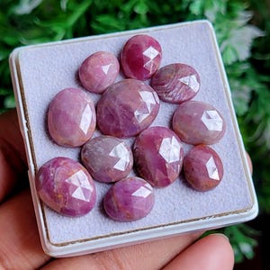 Puede incluir: Colección de gemas rosas facetadas, de forma ovalada y redonda. Las gemas se muestran en una bandeja blanca con interior gris claro. Las gemas tienen una superficie pulida y reflectante.