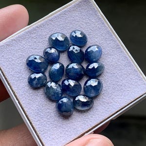 Puede incluir: Una colección de trece zafiros azules facetados de forma ovalada dispuestos en un grupo. Cada piedra preciosa muestra un tono azul profundo y saturado con una superficie pulida de múltiples facetas que refleja la luz sobre una almohadilla de terciopelo gris.