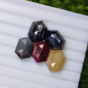 Puede incluir: Una colección de cinco piedras preciosas sueltas y facetadas de forma hexagonal. Las piedras incluyen dos azul oscuro, una gris, una rojo intenso y una amarilla translúcida, todas con superficies planas y pulidas con facetas.