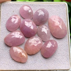 May include: A collection of ten oval-shaped, faceted pink gemstones arranged on a light gray textured surface. Each stone features a rose-colored hue with a polished, geometric cut that reflects light across its surface, showcasing various shades of pink.