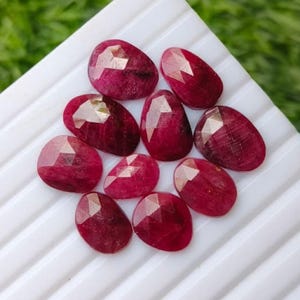 Puede incluir: Una colección de diez rubíes sueltos, facetados y de forma ovalada en un tono rojo profundo y saturado. Cada piedra preciosa presenta una superficie pulida de múltiples facetas que refleja la luz, mostrando una calidad translúcida y variaciones naturales.