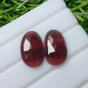 Könnte beinhalten: Zwei ovale, facettierte Edelsteine in einem tiefen rötlichen Rosaton. Jeder Stein hat eine polierte Oberfläche mit mehreren flachen, geometrischen Facetten, die das Licht reflektieren und eine durchscheinende Qualität sowie abgerundete Kanten aufweisen.